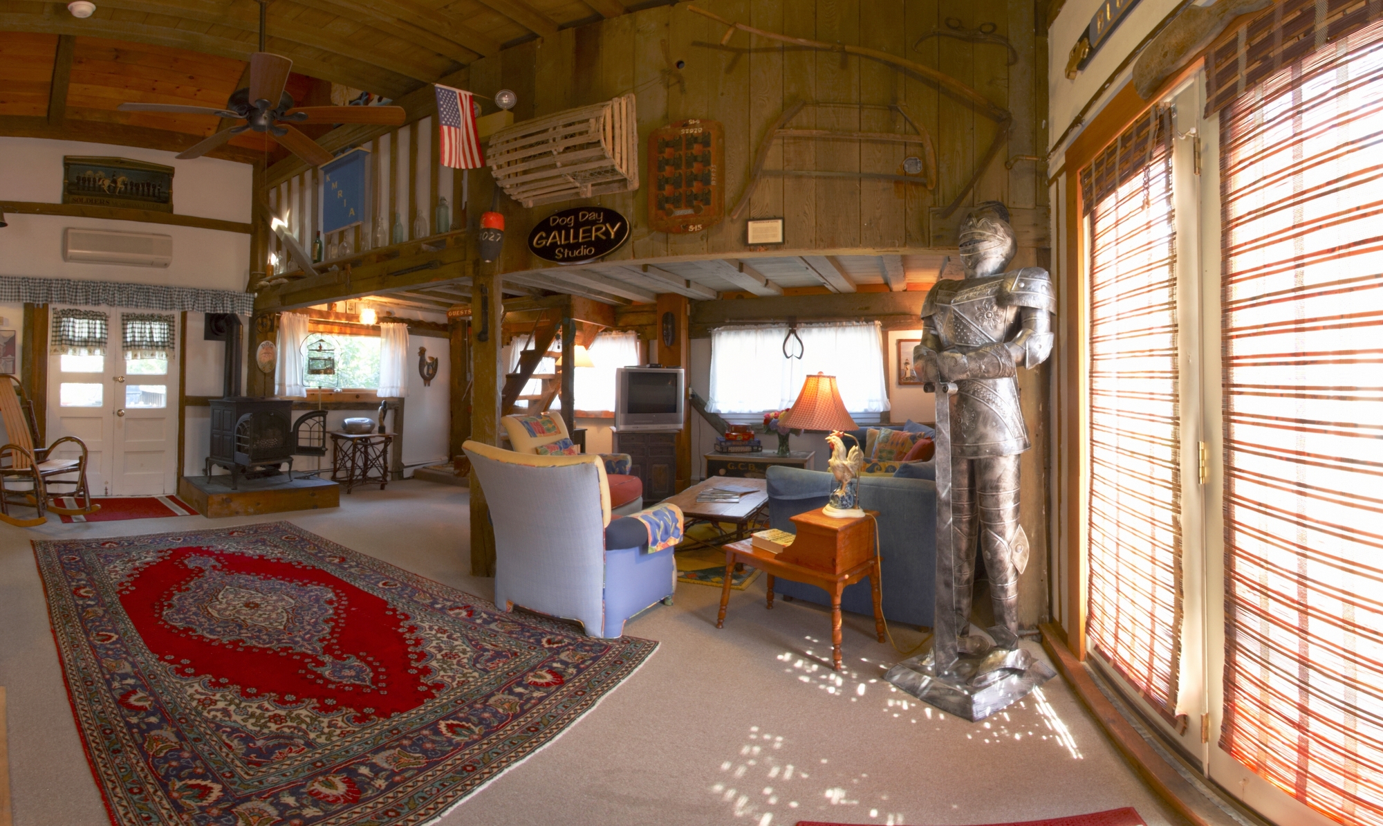 A photo of the Barn living room area. There is a red colored Oriental rug, a chair and lamp in the background. A knight in a suit of armor guards the entrance doors. A flag and old tools adorn the wall.