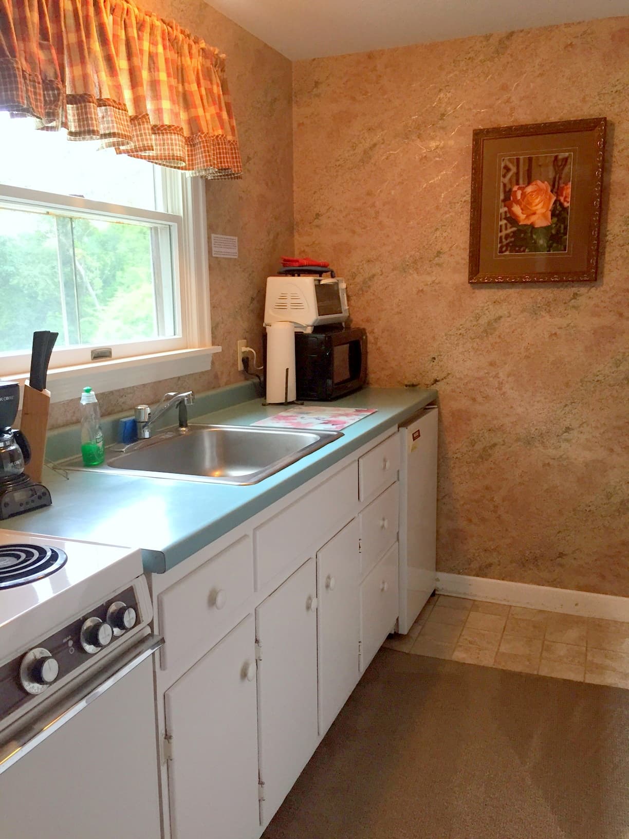 A small kitchen with white cabinets a green counter top and light streaming through the window. There is a framed photo of a rose.