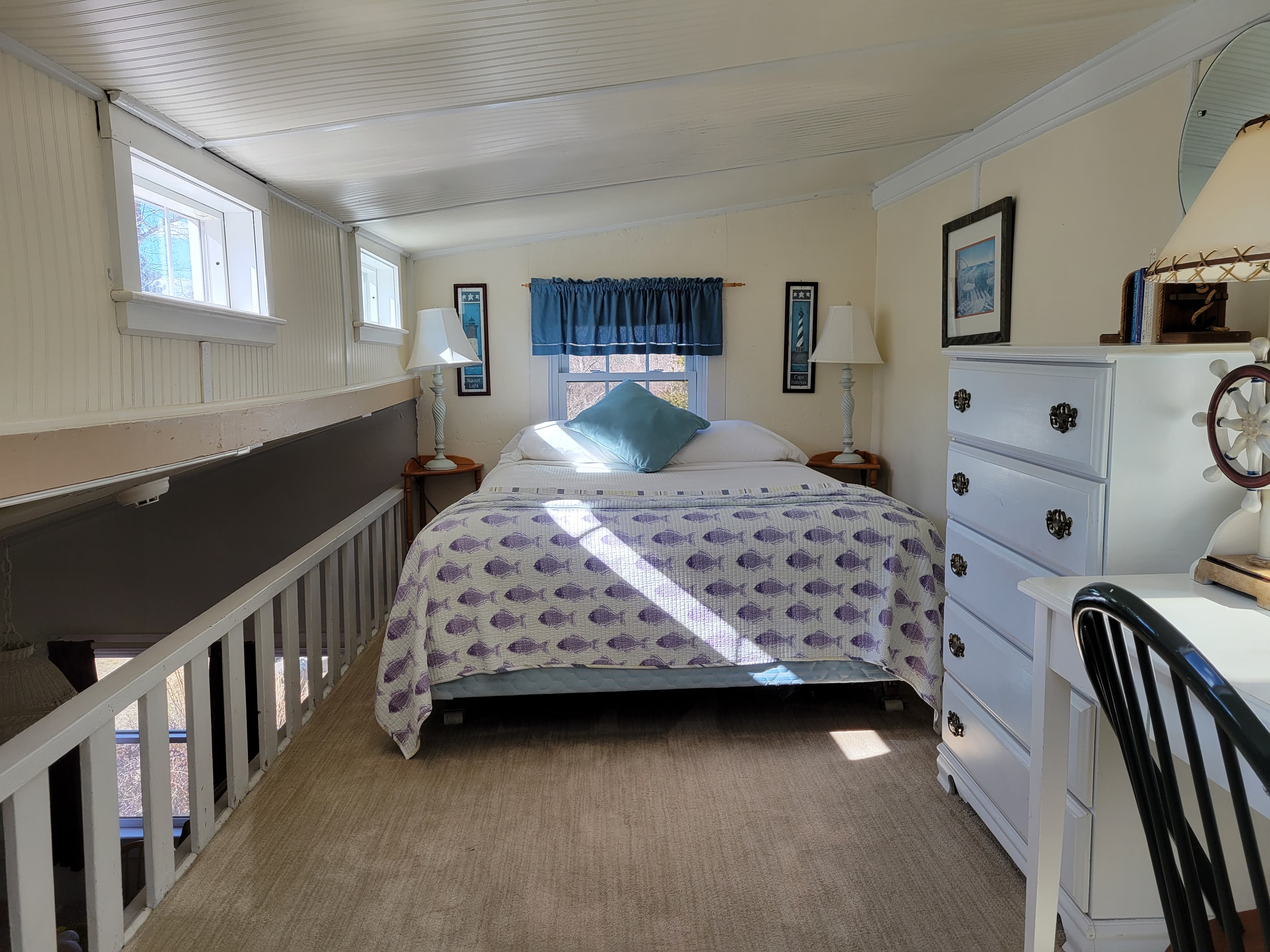 Loft bedroom bed of blues and whites with a white chest-of-drawers and light streaming in the small windows