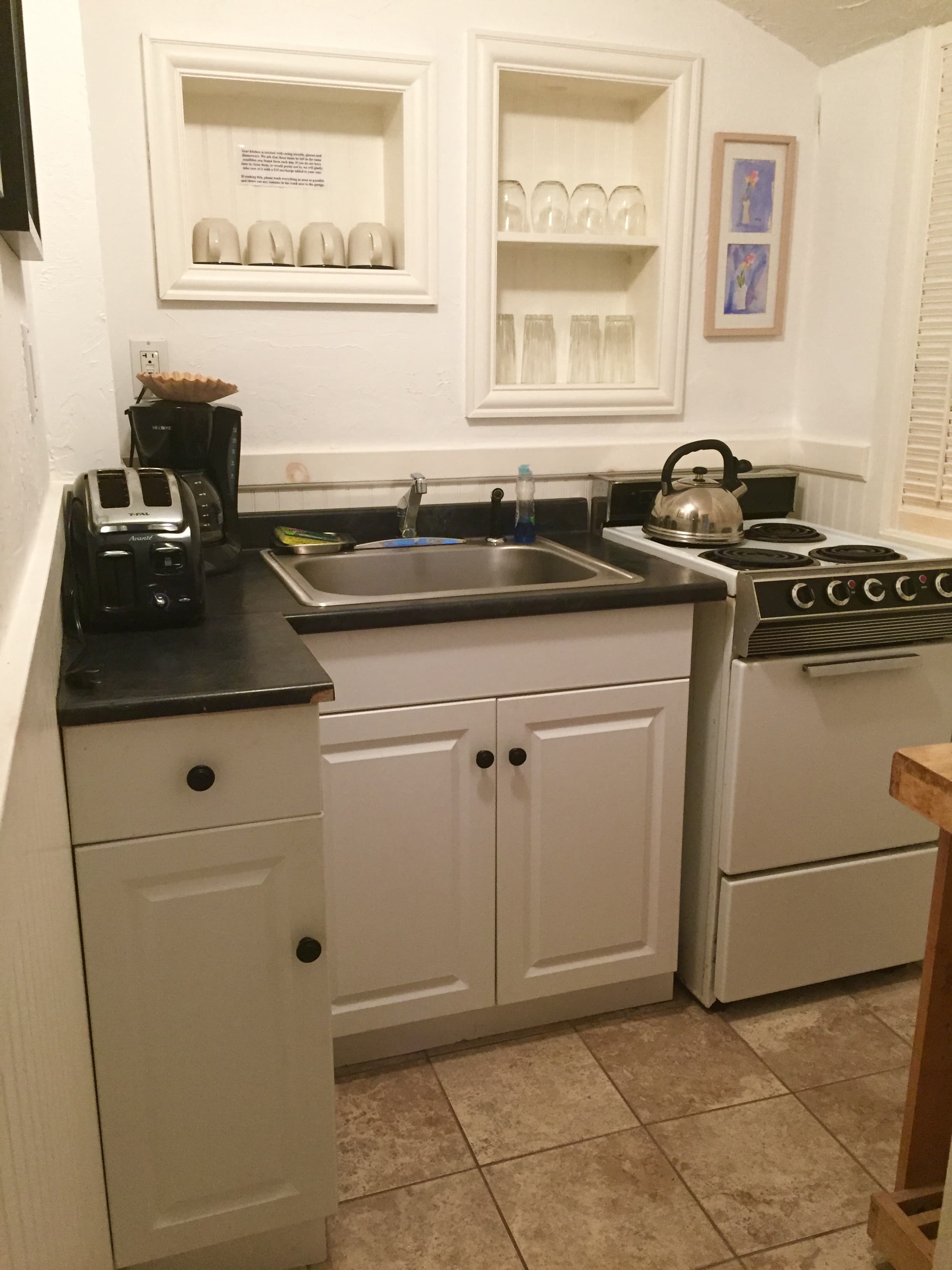 A white galley kitchen with black counter tops and built-in shelves with glasses on them