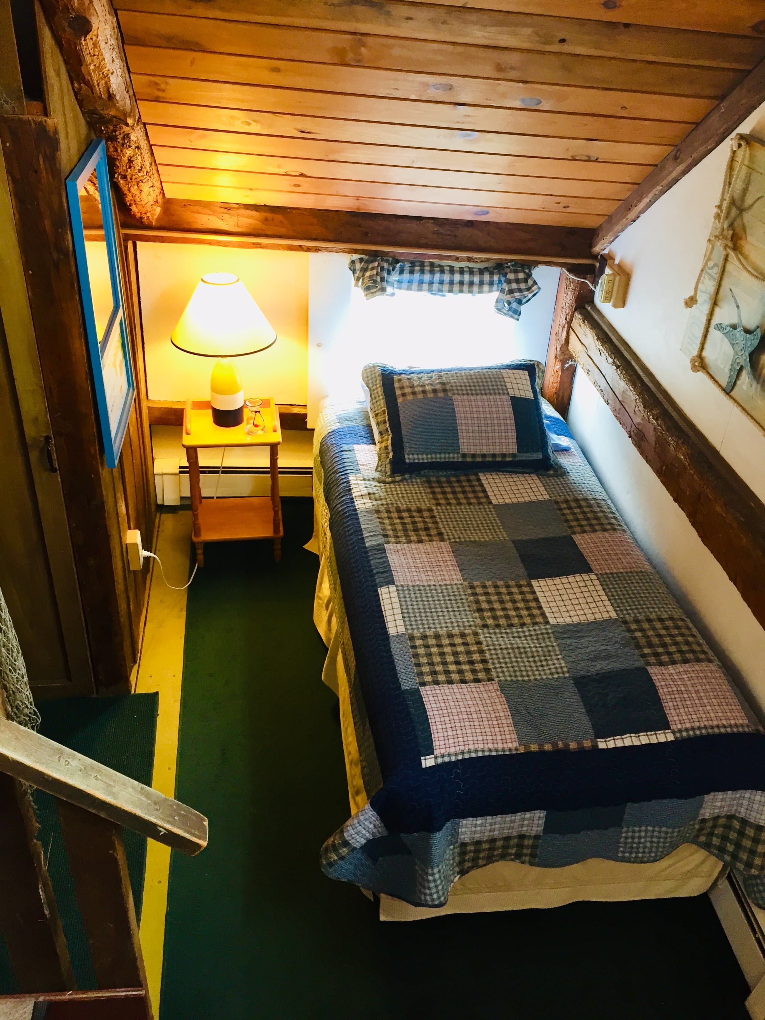 A twin bed in a loft with a blue quilt and a lamp on next to the bed.