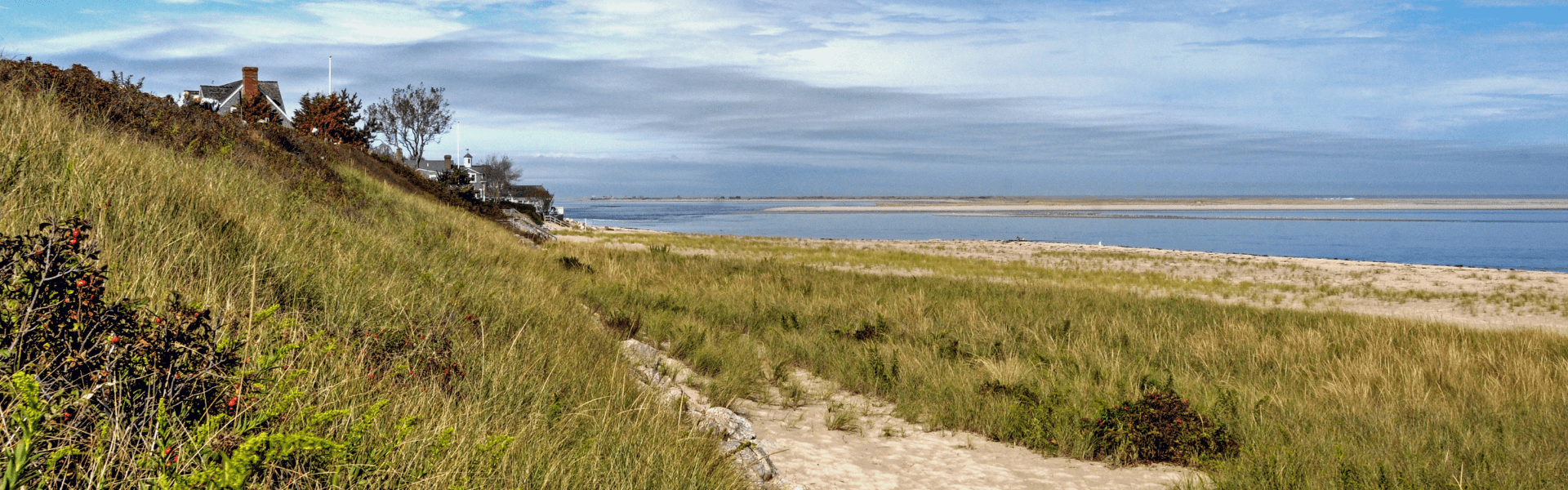 A coastal landscape featuring grassy dunes, a sandy beach, and a calm waterway under a cloudy sky.