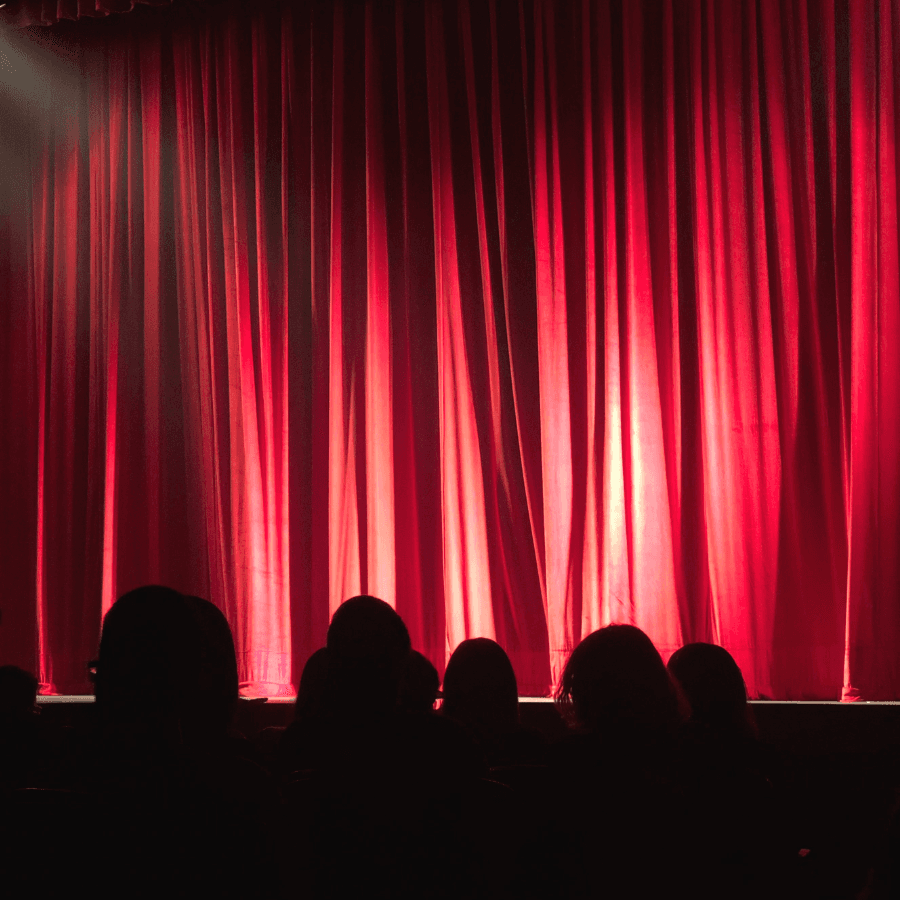A dimly lit theater stage with red curtains and silhouettes of an audience.